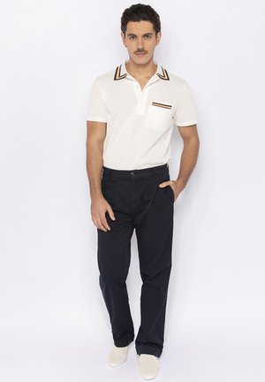 Witte poloshirt met contrasterende gestreepte kraag en zakdetail, gecombineerd met marineblauwe broeken en lichtgekleurde instapschoenen.