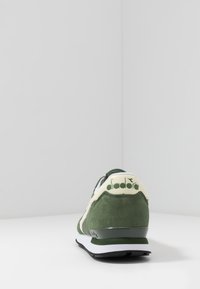 Green suede sneaker with a white midsole, featuring a cream logo on the heel, and a rounded shape with a textured upper.