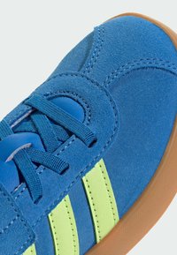 Blauwe suède sneakers met drie groene strepen, ronde veters en een tan rubberen zool. Getextureerde bovenkant en accenten zorgen voor diepte en detail.