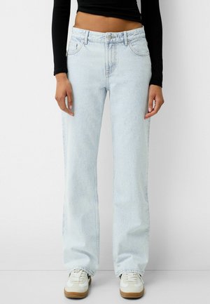 Person trägt hellblaue, gerade geschnittene Jeans, weiße Turnschuhe und ein schwarzes, langärmliges, kurzes Oberteil und steht vor einem schlichten Hintergrund.