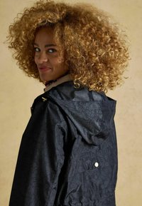 Giacca nera resistente all'acqua con motivo floreale, dotata di cappuccio, chiusura con cerniera e dettagli in metallo dorato. Tessuto strutturato.