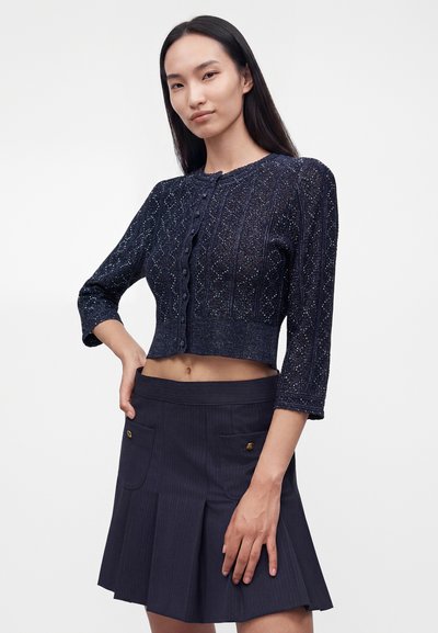 Femme aux longs cheveux foncés porte un cardigan court texturé bleu marine et une jupe plissée bleu marine avec des poches avant, debout avec une main sur la hanche.