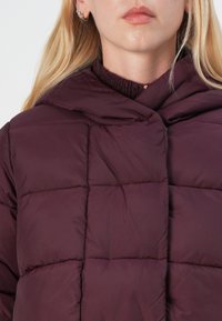 Giacca puffer bordeaux con motivo trapuntato e colletto alto. Texture liscia, chiusura frontale e accenni di un colletto interno più scuro.