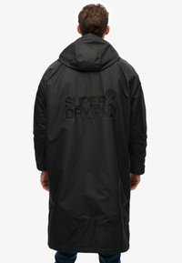 Zwarte waterdichte parka met capuchon, met een reliëf "SUPERDRYPN." logo op de achterkant, lange mouwen en een losse pasvorm.