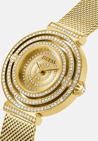 Zlatni sat s okruglim licem, spiralnim dizajnom i brojnim prozirnim kristalima koji okružuju brojčanik. Ima mrežastu narukvicu i teksturiranu krunicu.