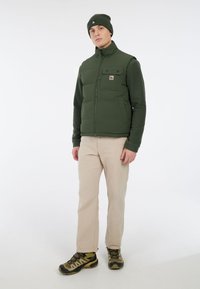 Man met groene beanie, groene mouwloze puffer vest over een olijfgroene trui, beigekleurige broek en groene wandelschoenen, staand tegen een witte achtergrond.