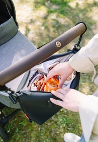 Handen plaatsen een oranje babyfles in een zwarte kinderwagenrugzak met kleding en een roze smartphone, buiten op het gras.