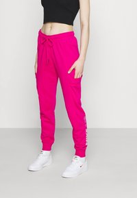 Růžové joggingové kalhoty s elastickým pasem na šňůrku, bočními kapsami a bílým textem "NIKE AIR" podél levé nohy. V kombinaci s bílými teniskami.