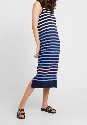 Kobieta w granatowej, bezrękawowej sukience midi z poziomymi białymi, niebieskimi i różowymi paskami, w czarnych sandałach z klamrą, stoi bokiem.