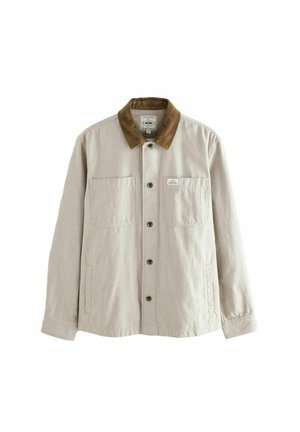 Man draagt een beige corduroy jas met een bruine kraag, staat buiten met zijn hand in zijn zak, bomen en een grasveld op de achtergrond.