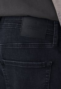 Vaqueros de mezclilla negra con una superficie texturizada. Presentan un bolsillo trasero y una etiqueta rectangular de cuero negro con texto en relieve.