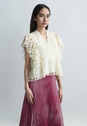 Τοπ από δαντέλα με κεντήματα λουλουδιών σε ελεφαντόδοντο, μανίκια με στρώσεις βολάν, συνδυασμένο με πλισέ φούστα ομπρέ σε αποχρώσεις του ροζ έως του βουργουνδίας.