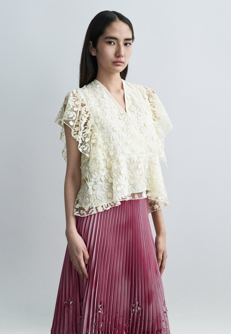 Τοπ από δαντέλα με κεντήματα λουλουδιών σε ελεφαντόδοντο, μανίκια με στρώσεις βολάν, συνδυασμένο με πλισέ φούστα ομπρέ σε αποχρώσεις του ροζ έως του βουργουνδίας.