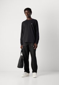 Ung person står med en svart totebag iförd svart langærmet skjorte, mørke jeans og hvite sneakers mot en hvit bakgrunn.