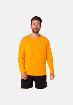Mann mit leuchtend orangefarbenem Langarmshirt und schwarzen Shorts, der vor einem einfarbigen hellen Hintergrund steht.