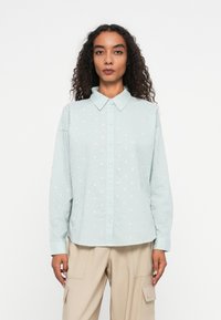 Light blue shirt made of textured fabric, featuring white polka dots, button-up front, and a pointed collar. Long sleeves with cuffs.