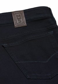 Dunkelblaue Jeans aus Denim mit einem braunen Lederpatch und geprägtem "M5"-Logo. Der schwarze Faden hebt sich vom Stoff ab. Die Detailgestaltung der Taschen ist sichtbar.