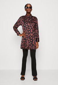 Juodo ir raudono satino tunika su leopardų raštu, ilgomis rankovėmis, mygtukų uždarymu, derinama su juodomis plačiomis kelnėmis ir juodais šlepetėmis.