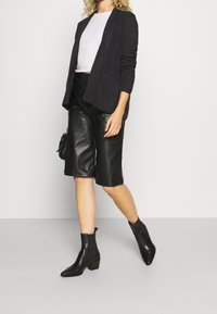 Mörkgrå öppen front cardigan, vit t-shirt, svarta lädershorts och svarta spetsiga ankelskor. Släta texturer och ett strukturerat passform.