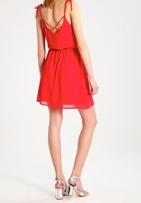 Rotes ärmelloses Kleid mit tailliertem Schnitt, gerafftem Rock und überkreuzten Riemen im Rücken. Getragen mit metallischen Absatzsandalen.