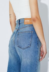 High-waisted blue denim jeans with a relaxed fit, featuring a brown brand label on the back and two back pockets with contrasting stitching.
