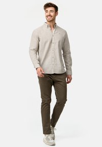 Uomo sorridente che cammina, indossa una camicia beige con bottoni, pantaloni verde oliva e sneaker grigie su uno sfondo bianco uniforme.