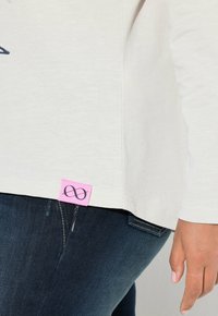 Helles langärmliges Hemd mit einem seitlichen Logo-Tag in Pink, das ein Unendlichkeitssymbol zeigt. Kombiniert mit dunkelblauen Jeans.