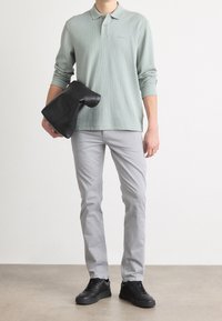 Długi rękaw zielonej polówki, szare spodnie i czarne trampki. Trzyma czarną skórzaną torbę. Prosty design o luźnym kroju.