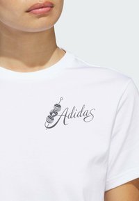 Bílé bavlněné tričko s černou grafikou činky a nápisem "Adidas" v kurzívním písmu, s kulatým výstřihem a krátkými rukávy.