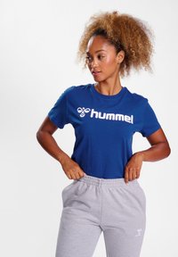 Blå bomulds t-shirt med hvid "hummel" logo, parret med lysegrå sweatpants med snøre. Glat tekstur, korte ærmer, afslappet pasform.