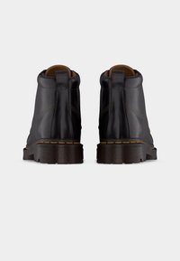 Ζευγάρι μαύρα δερμάτινα μποτάκια αστραγάλου με παχύ λαστιχένιο πέλμα και κίτρινη ραφή, φωτογραφημένα από πίσω σε απλό φόντο.