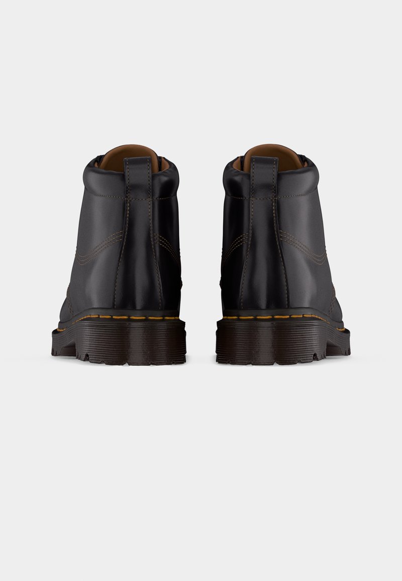 Een paar zwarte leren enkellaarsjes met dikke rubberen zolen en gele stiksels, gezien vanaf de achterkant tegen een effen achtergrond.