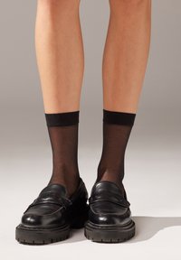 Mocassini neri con suola spessa e finitura liscia, abbinati a calzini alla caviglia neri trasparenti con una banda solida nella parte superiore.