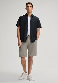 Camicia a maniche corte nera con bottoni sopra una maglietta bianca, abbinata a pantaloni corti khaki con trama e sneakers bianche, in piedi davanti a uno sfondo neutro.