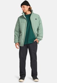 Giacca imbottita verde mentolato con zip frontale, tasche laterali e colletto alto. Abbinata a una maglietta verde scuro e pantaloni neri.