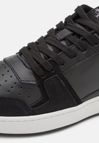 Zapatilla negra que combina cuero liso y gamuza, con puntera redonda, detalles perforados y cordones planos negros sobre una suela de goma blanca.