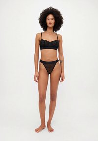 Ensemble de lingerie noire comprenant un bralette en tissu transparent à motif de pois et une culotte taille haute assortie avec un design similaire.