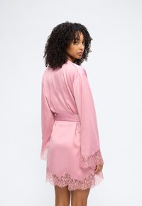 Γυναίκα με σγουρά μαλλιά που φοράει ροζ σατέν ρόμπα με δαντέλα στα μανίκια και στο τελείωμα, κοιτάζει πάνω από τον ώμο της, μπροστά σε ένα απλό φόντο.