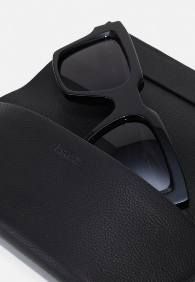 Μαύρα γυαλιά ηλίου με γωνιακούς σκελετούς τοποθετημένα σε μια υφασμάτινη μαύρη θήκη. Και τα δύο αντικείμενα διαθέτουν κομψό, ματ φινίρισμα.