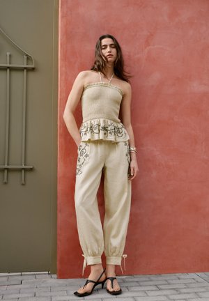 Žena u bež topu bez naramenica s volanima i ukrašenim bež hlačama s crnim sandalama s potpeticama stoji uz crveni zid.