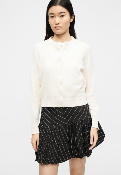 Νέα γυναίκα με μακριά σκούρα μαλλιά, που φοράει μια κρεμ ζακέτα με κουμπιά και μια μαύρη πλισέ φούστα με διαγώνιες λευκές ρίγες.
