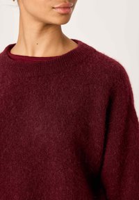 Gros plan d'une personne portant un pull doux et pelucheux de couleur rouge foncé avec un col rond, sur un fond clair.