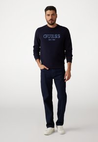 Granatowy sweter z napisem "GUESS EST. 1981" wyszytym na niebiesko, w zestawie z ciemnoniebieskimi dżinsami i białymi trampkami. Tkanina ma gładką fakturę.