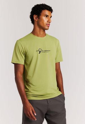 Man in een lichtgroen "Protest" T-shirt en donkere broek, staand met één hand in de zak, kijkend naar rechts tegen een effen achtergrond.