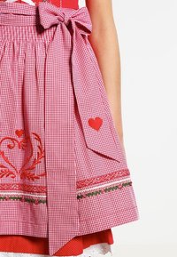 Tablier en vichy rouge et blanc avec des cœurs brodés, des bordures fleuries et un grand nœud noué à la taille sur une robe rouge avec un galon en dentelle blanche.