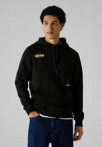 Sudadera con capucha negra y acentos del logotipo amarillos, con bolsillo frontal y cordón. Presenta una textura de tela y un ajuste relajado.