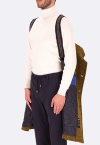 Cappotto imbottito nero con accenti giallo-marroni, indossato sopra un dolcevita bianco. Caratterizzato da cinturini regolabili neri con testo blu. Pantaloni blu navy.