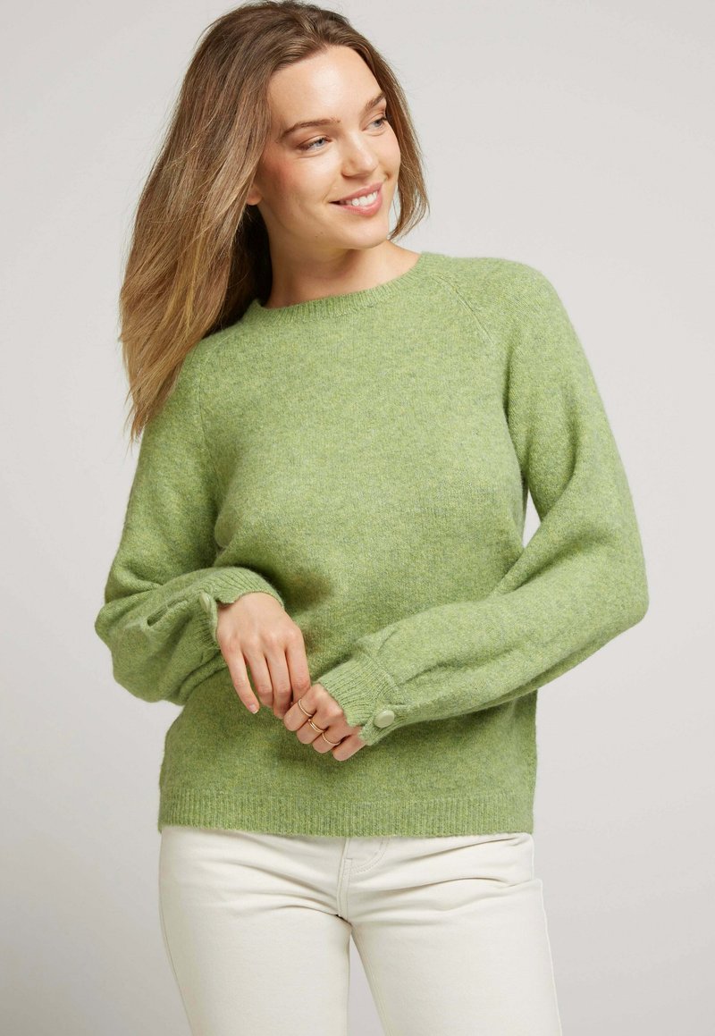Pull en tricot vert avec un col rond, des manches raglan et des accents à boutons sur les poignets. Texture douce et coupe décontractée.