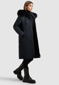 Parka nera lunga con cappuccio in pelliccia sintetica, dotata di chiusura lampo frontale, due tasche laterali e una finitura leggermente lucida. Vengono indossate scarpe stivali nere con lacci.