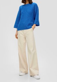 Persona che indossa un ampio maglione blu lavorato a maglia, pantaloni beige larghi e scarpe da ginnastica bianche, in piedi con una mano in tasca.